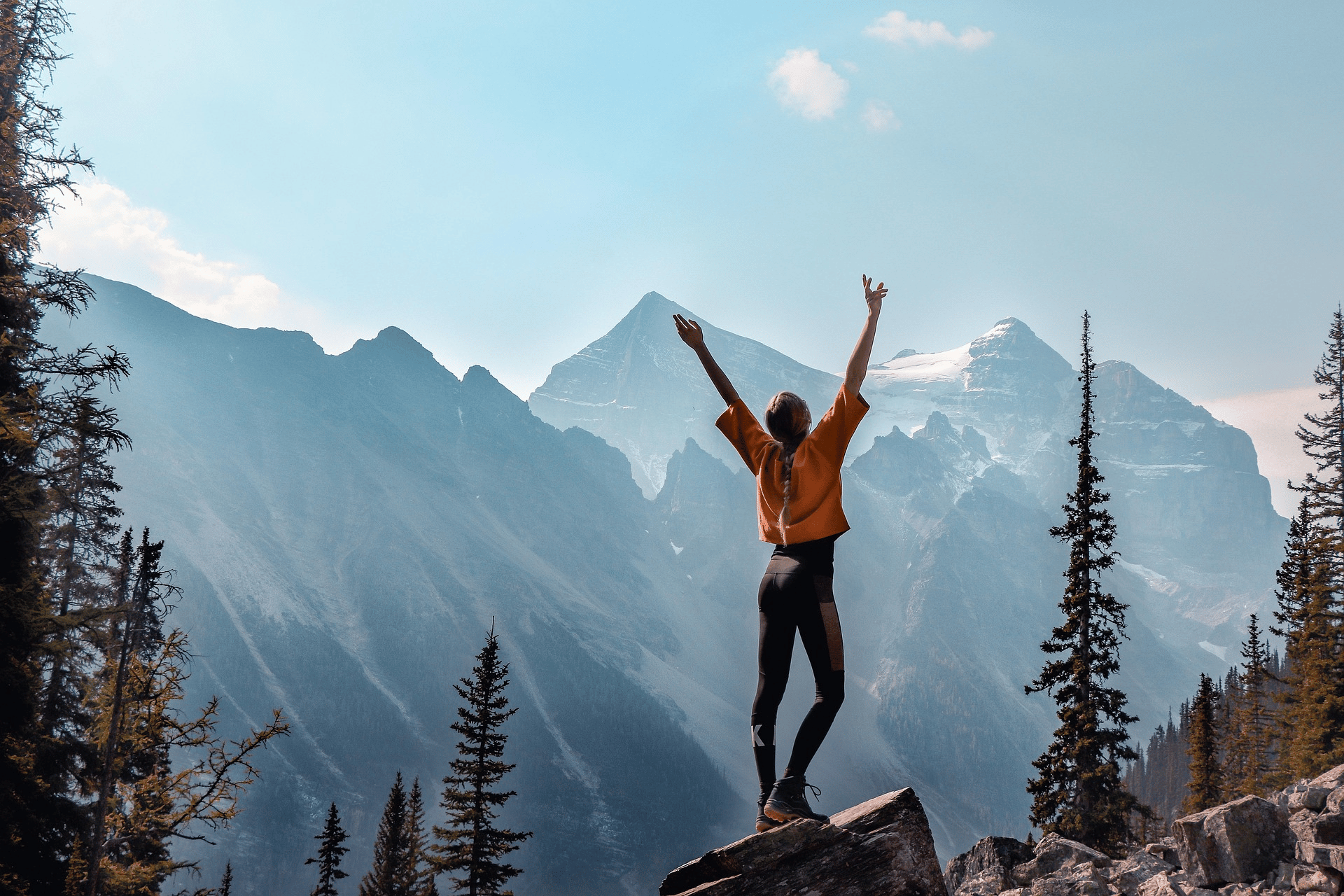 Femme en randonnée debout sur un rocher, levant les bras face à un paysage de montagnes majestueuses avec des sapins. Équipement outdoor adapté à l’aventure.